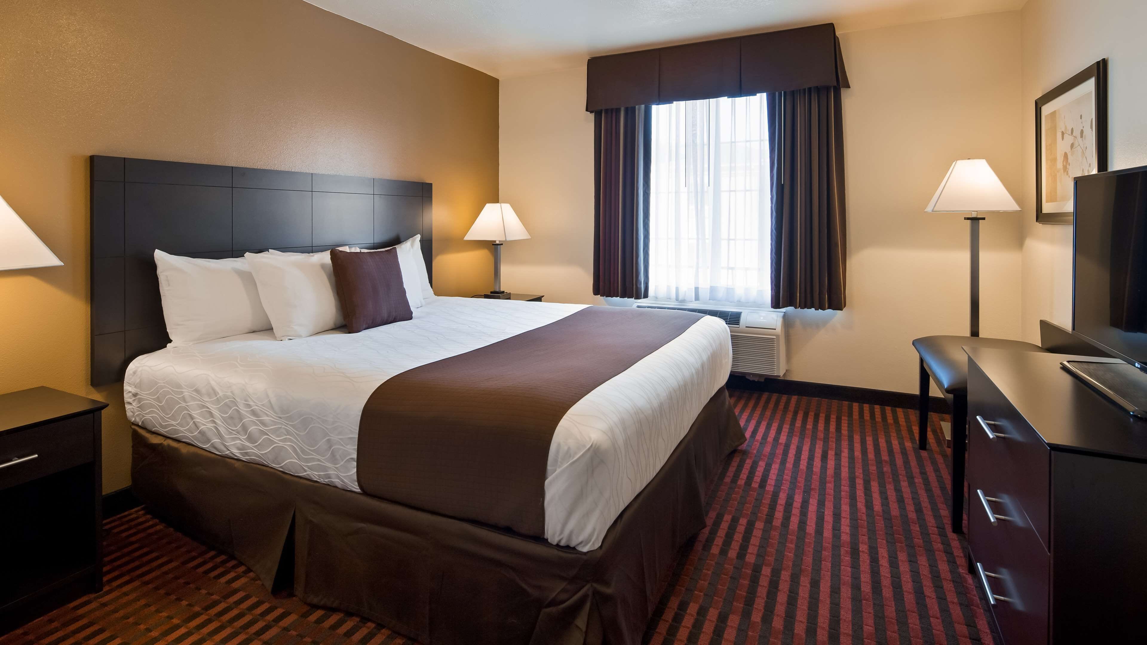 A hotel room with a large bed, two bedside tables, a desk with a chair, a window with curtains, and a wall-mounted TV.