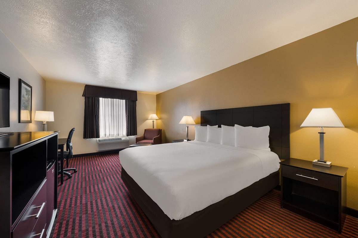 A hotel room with a large bed, two bedside tables with lamps, a desk with a chair, and a window with curtains.