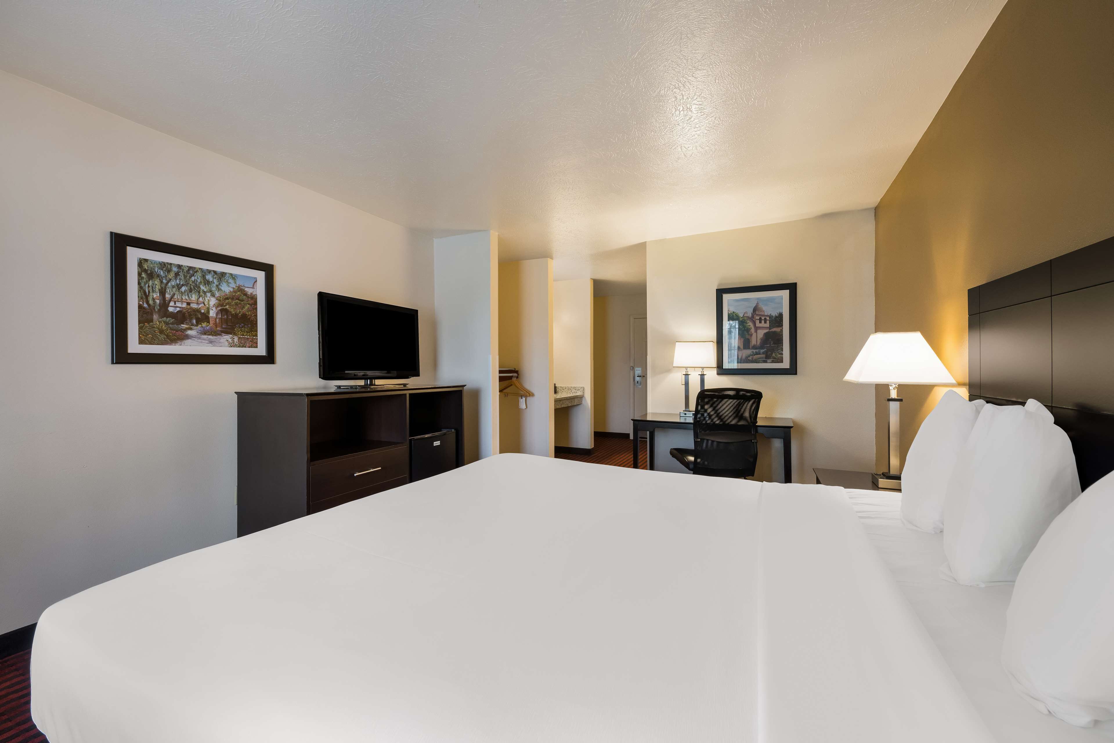 Une chambre d’hôtel avec un grand lit, un bureau avec une chaise, une télévision, une lampe et deux tableaux encadrés accrochés aux murs.