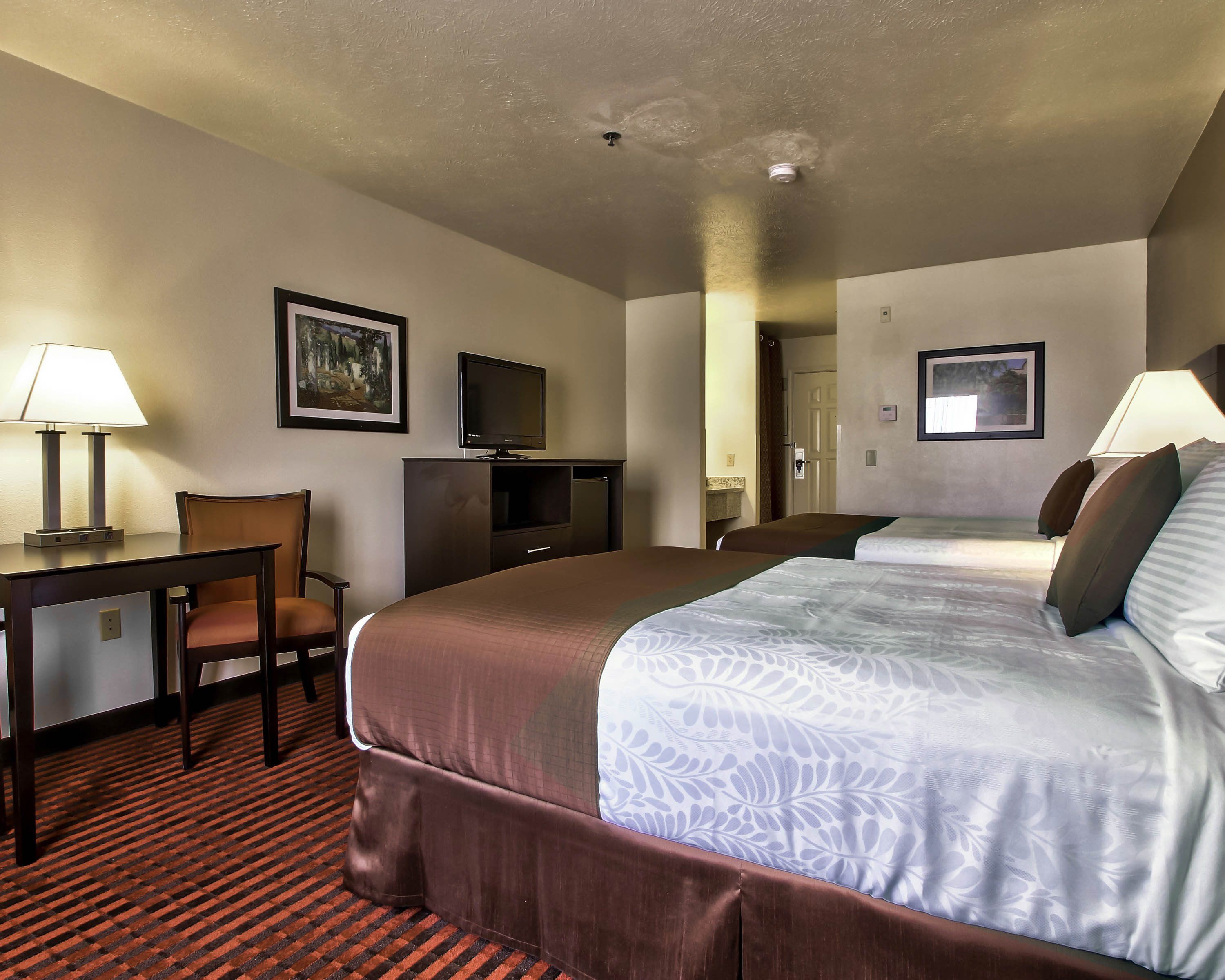 A hotel room with a large bed, a desk with a lamp, a television, and framed artwork on the walls.