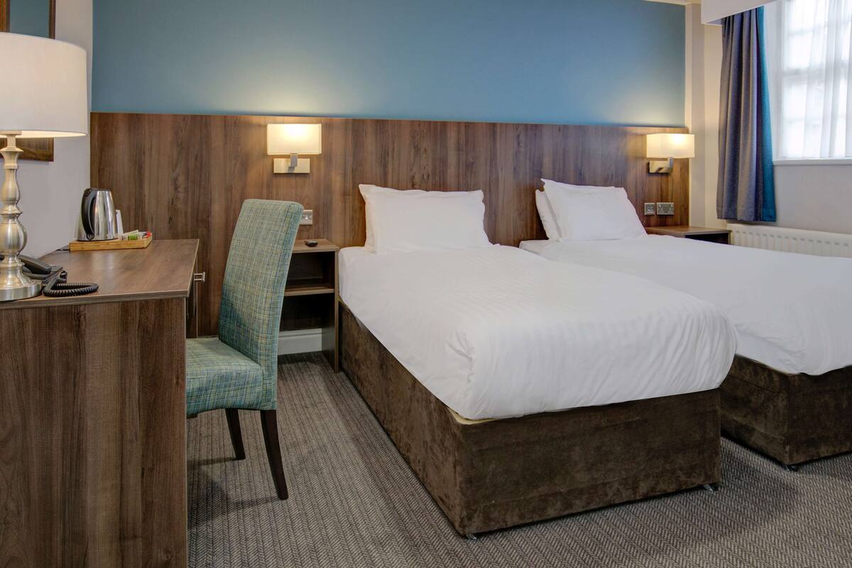 A hotel room with two beds, a desk, a chair, and a lamp.