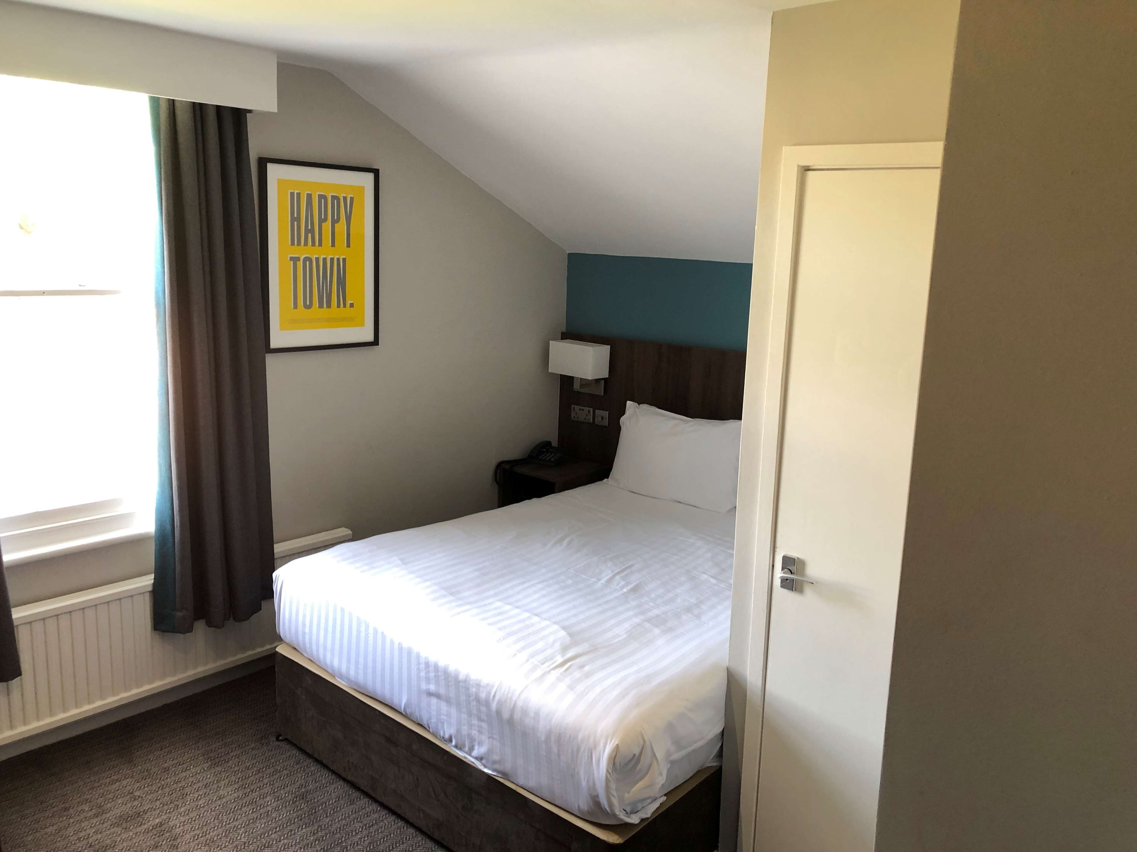 A hotel room with a bed, a nightstand, a lamp, a framed 'HAPPY TOWN' sign, a window with curtains, and a door.