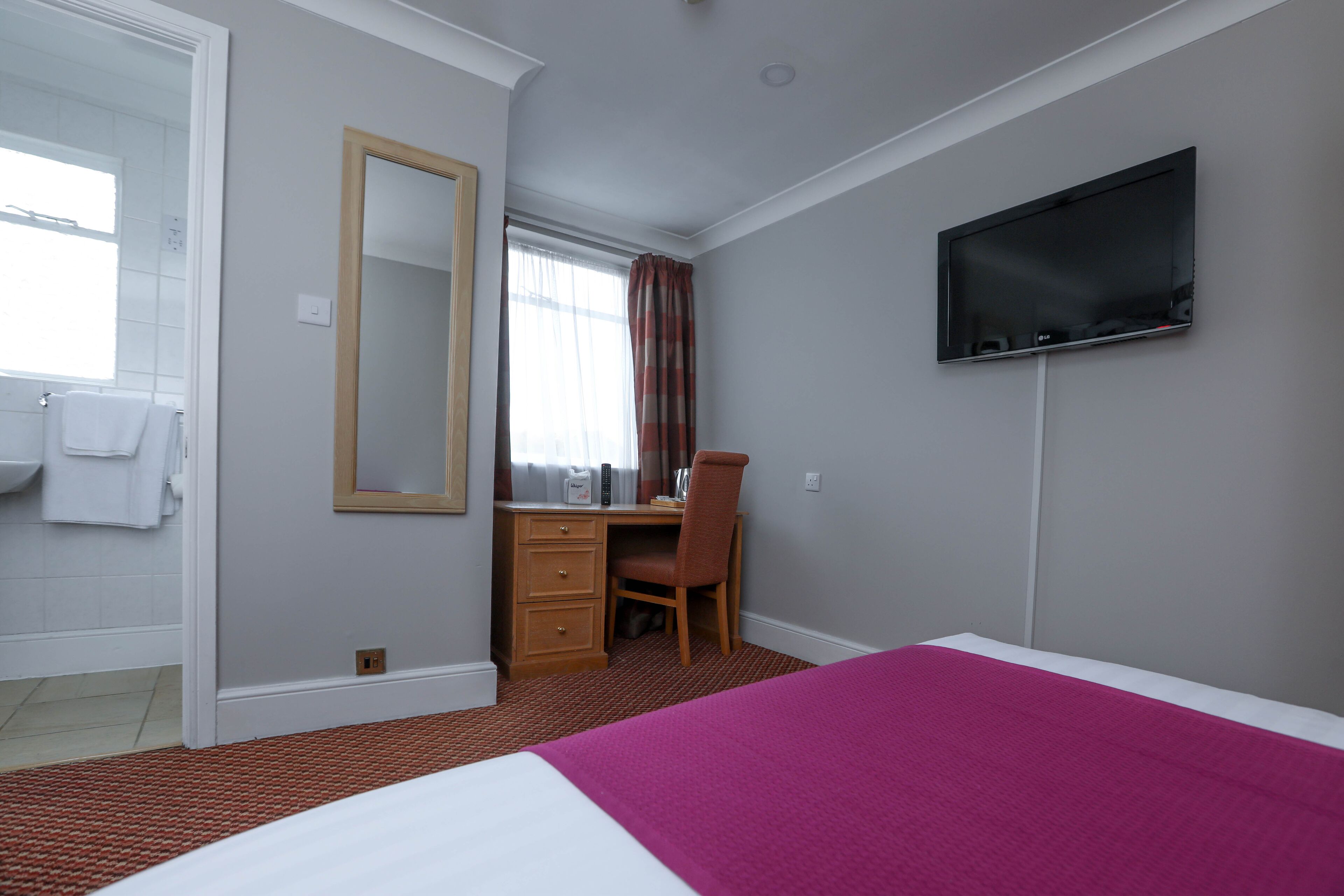 Habitación de hotel con cama, escritorio, silla, televisión y baño con lavamanos y espejo.