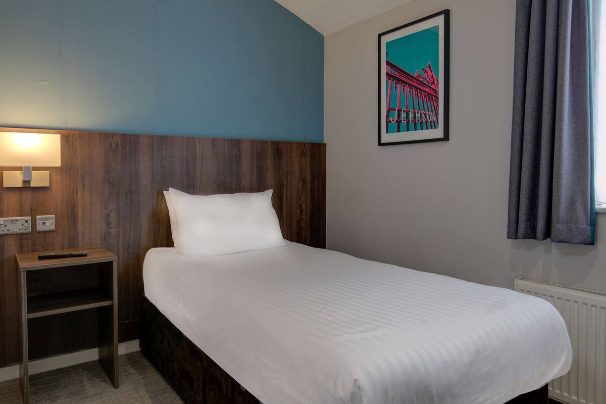 A hotel room with a single bed, a nightstand, a wall-mounted lamp, and a framed picture of a bridge.