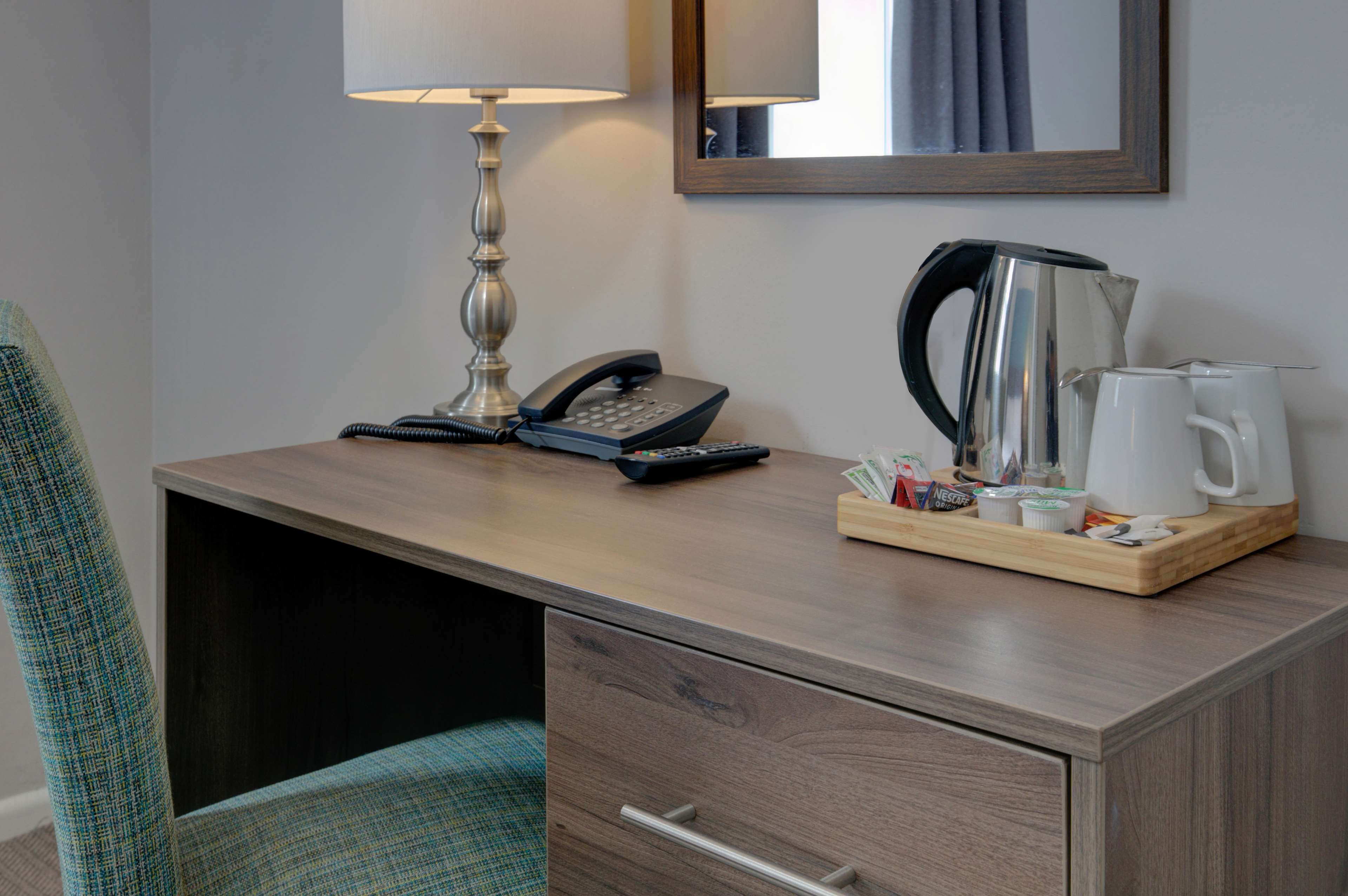A hotel room with a wooden desk, a lamp, a telephone, a coffee maker, and a tray with various items.