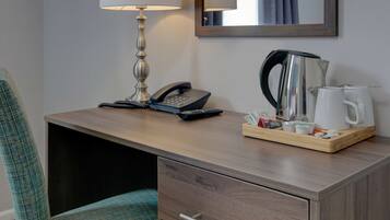 A hotel room with a wooden desk, a lamp, a telephone, a coffee maker, and a tray with various items.