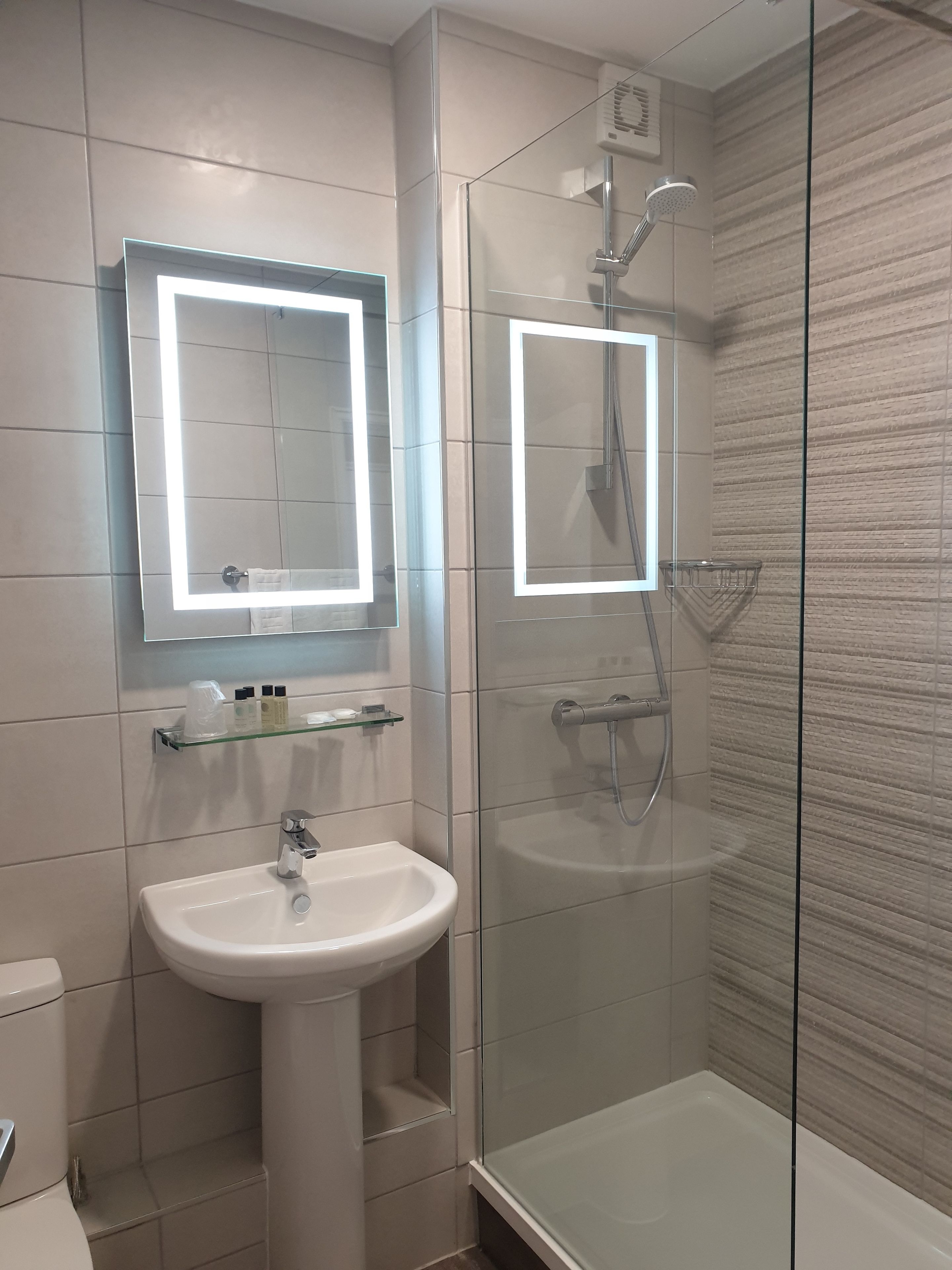 Un bany modern amb un envàs de dutxa de vidre, un lavabo de pedestal i un mirall amb il·luminació integrada.