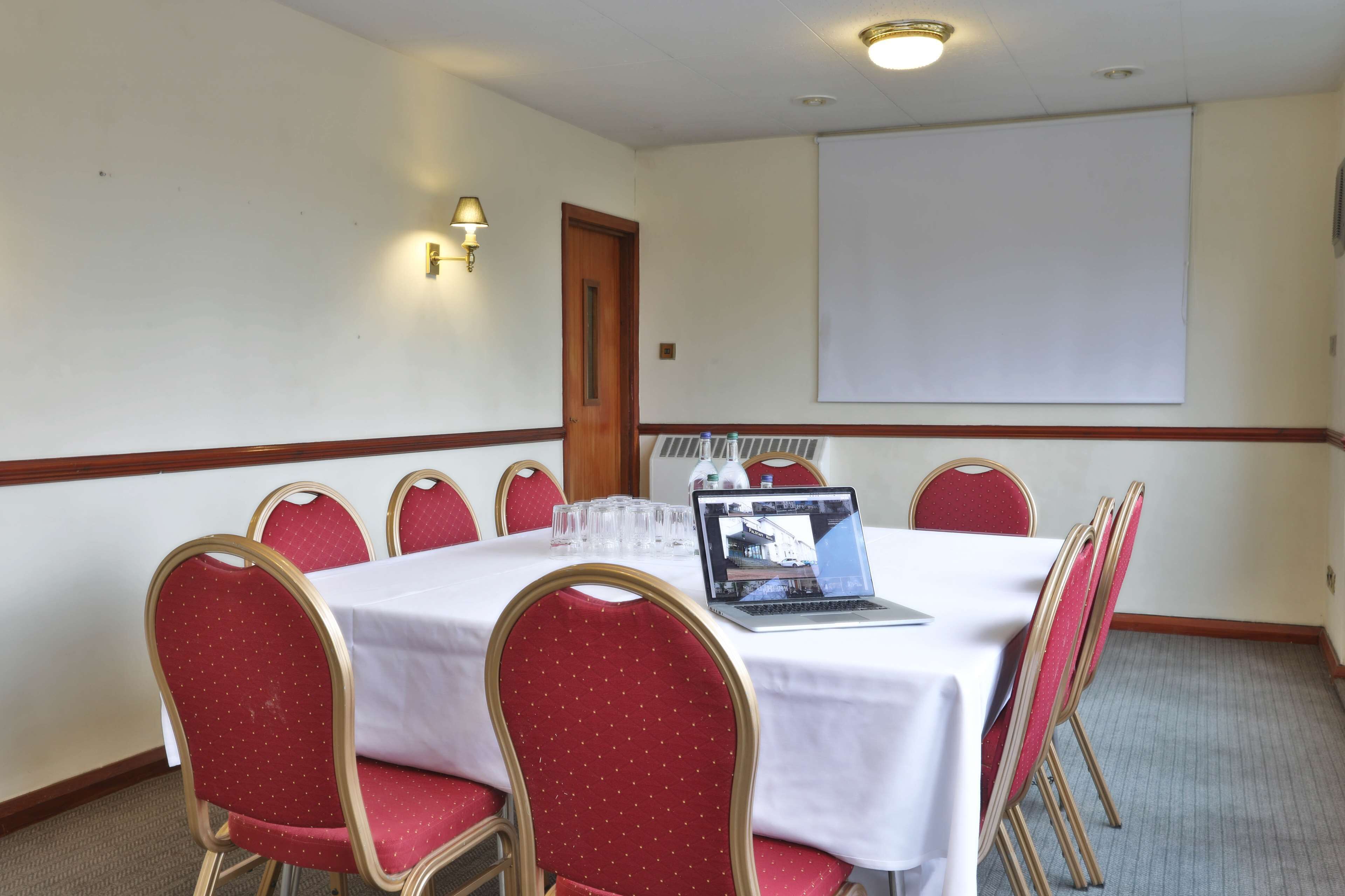 A conference room with a white table, red chairs, a laptop, and a projector screen.