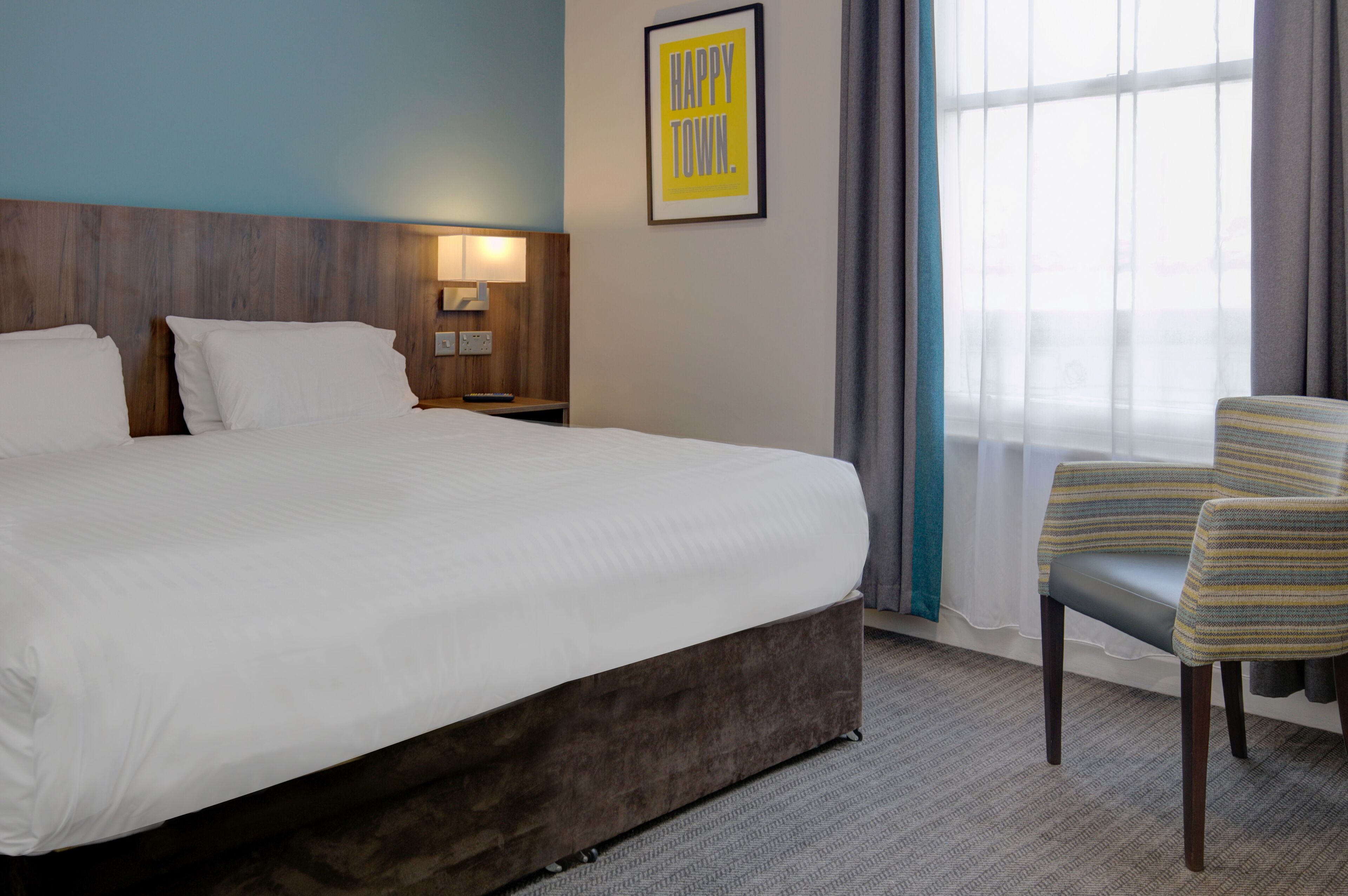A hotel room with a large bed, a nightstand with a lamp, a chair, and a window with curtains.