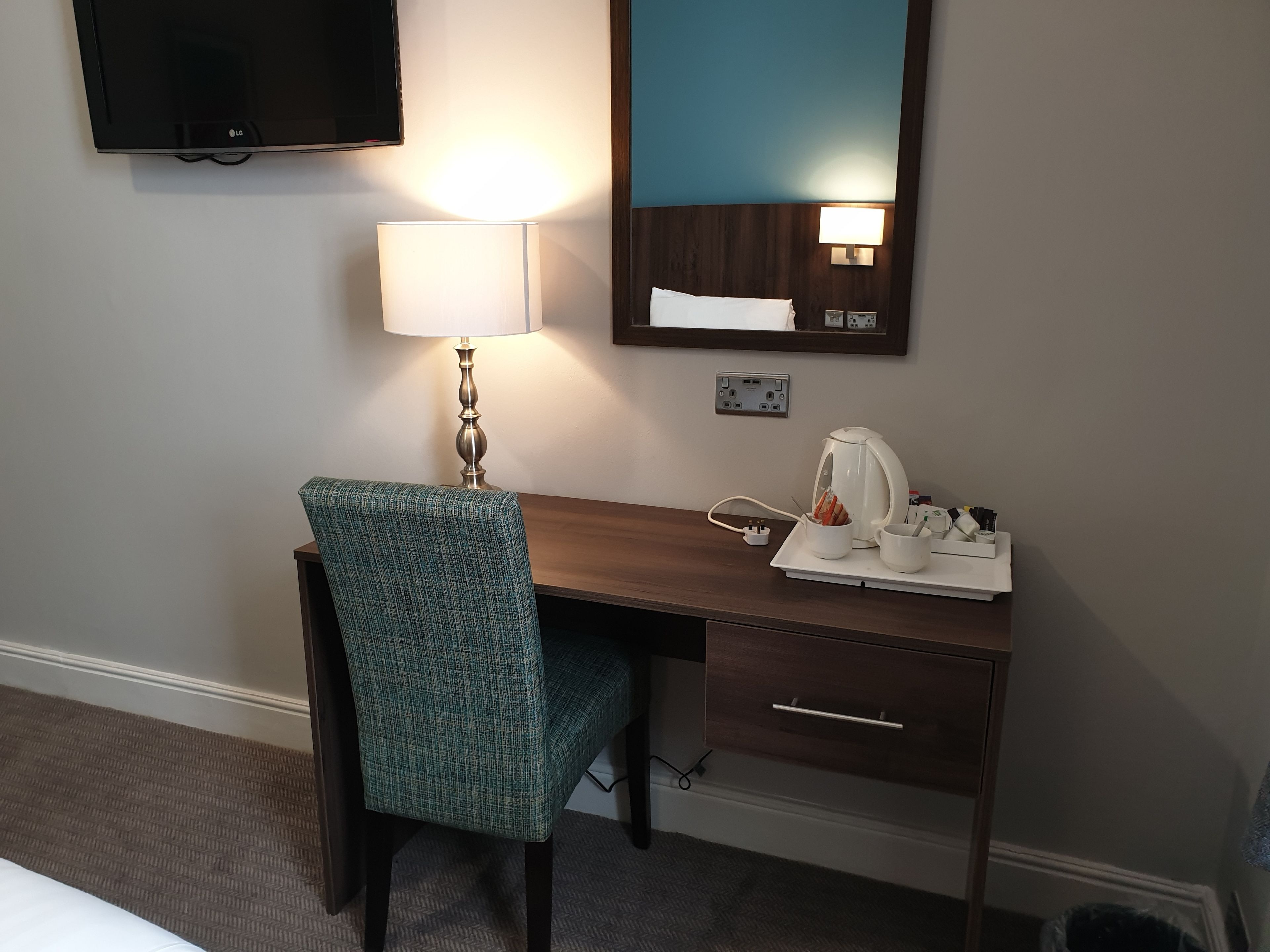 Ein Hotelzimmer mit Schreibtisch, Stuhl, Lampe, Spiegel, Fernseher und Kaffeemaschine.