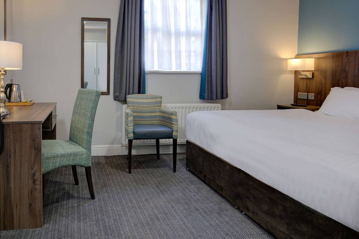 A hotel room with a bed, a chair, a desk with a lamp, a radiator, and a window with curtains.