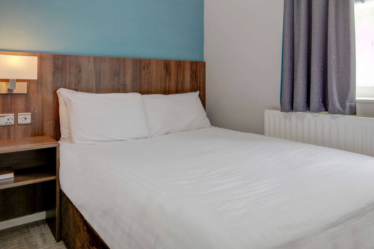A hotel room with a wooden headboard, a white bedspread, a nightstand with a lamp, and a window with curtains.