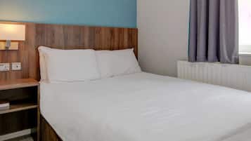 Una habitación de hotel con cabecera de madera, cubrecama blanco, mesita de noche con lámpara y ventana con cortinas.