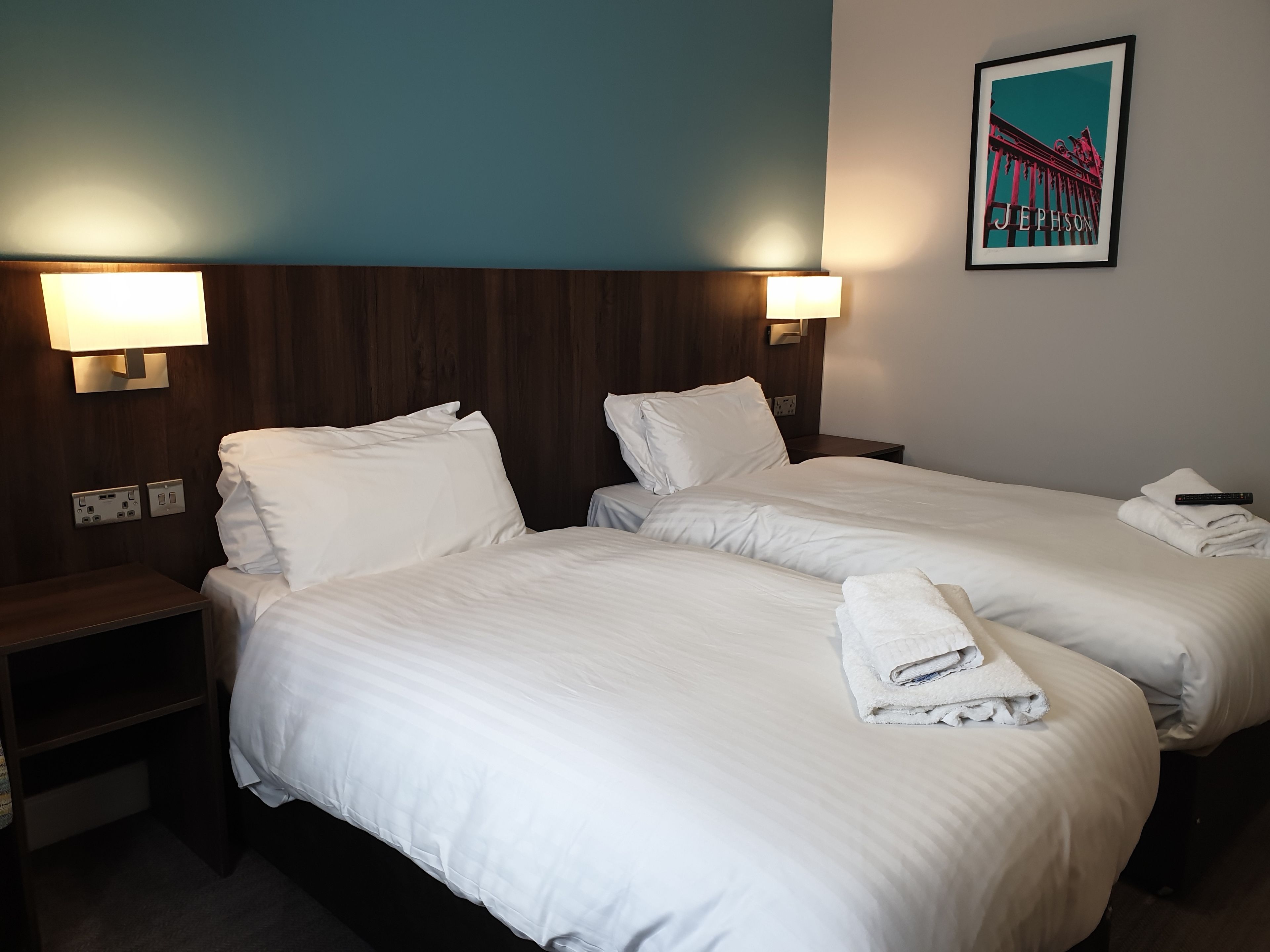 A hotel room with two beds, each with white linens and pillows, a nightstand with a lamp, and a wall-mounted light fixture.