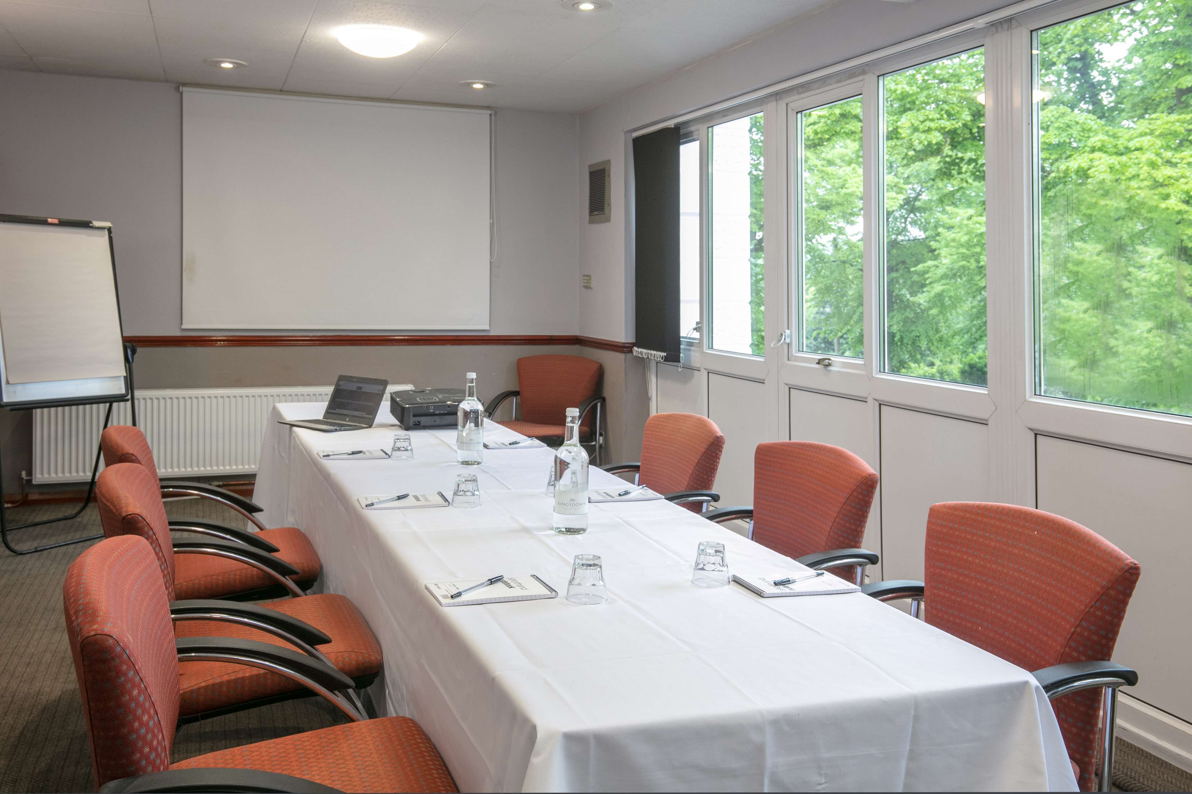 A conference room with a long table, chairs, a projector screen, and a window with a view of trees.