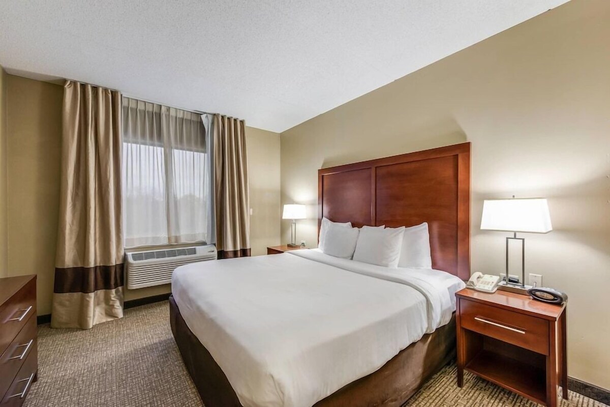 A hotel room with a large bed, two bedside tables with lamps, a wooden headboard, a window with curtains, and a wall-mounted air conditioning unit.