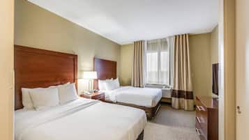 A hotel room with two beds, a wooden headboard, a bedside table with a lamp, a TV, and a window with curtains.
