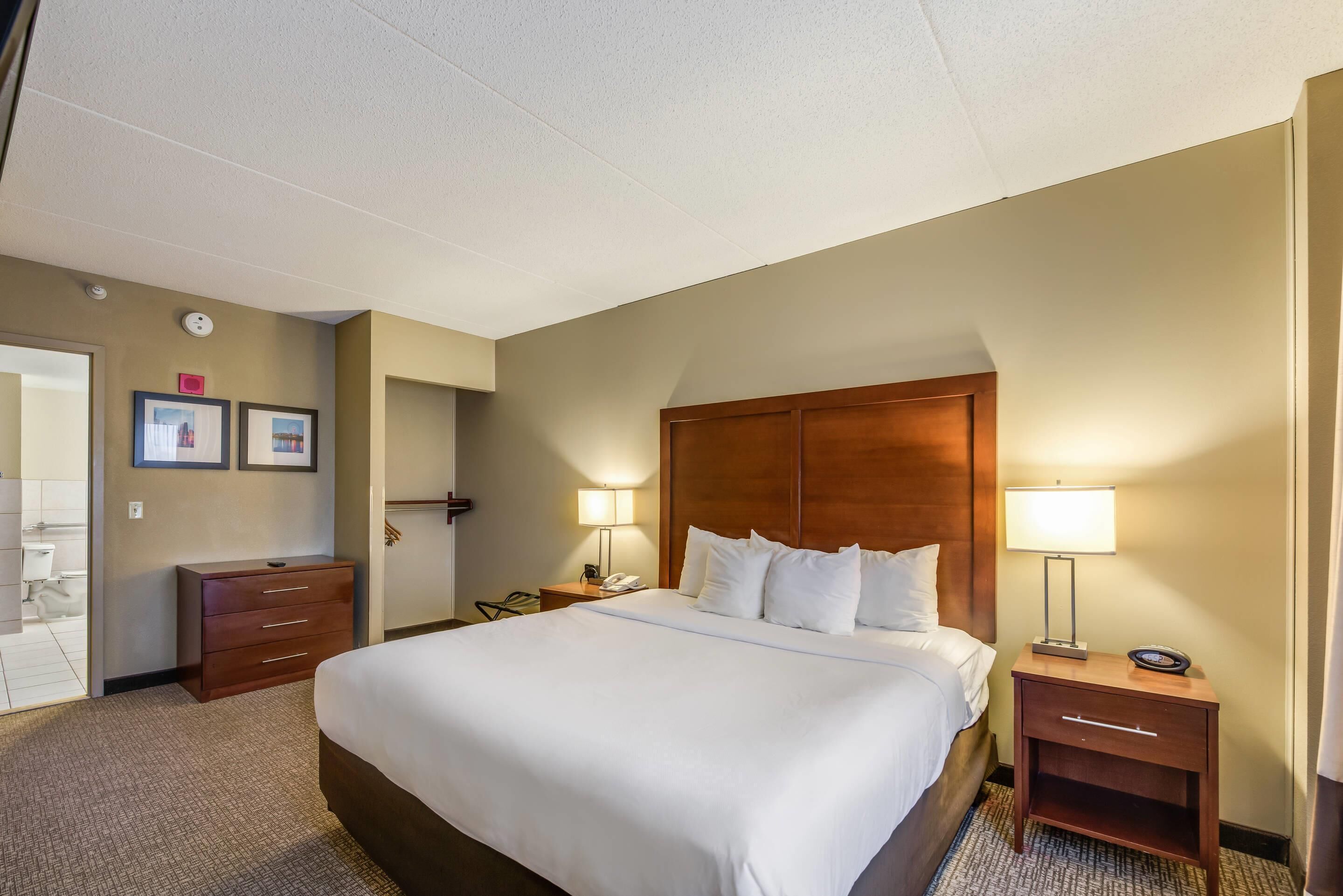 A hotel room with a large bed, two bedside tables with lamps, a wooden headboard, and a door leading to a bathroom.