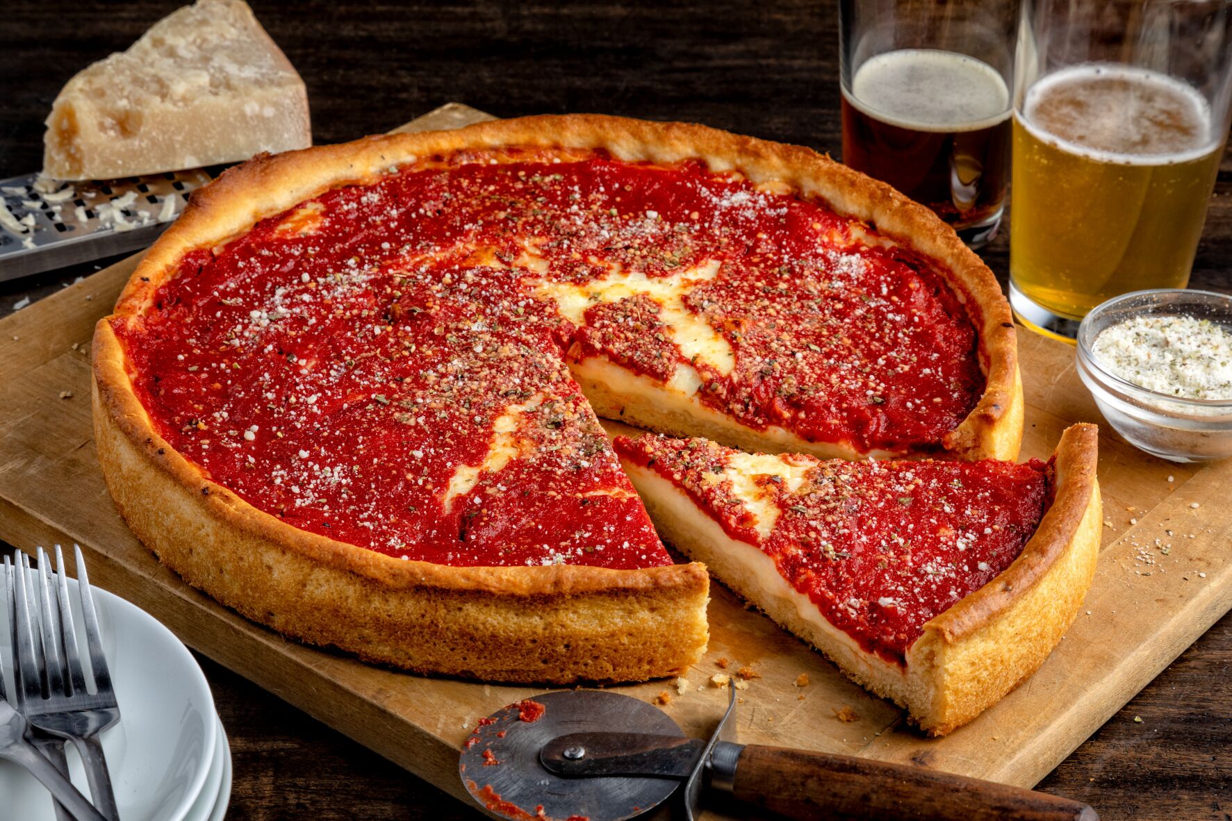 A deep-dish pizza with a slice missing, a beer glass, and a cheese grater on a wooden board.