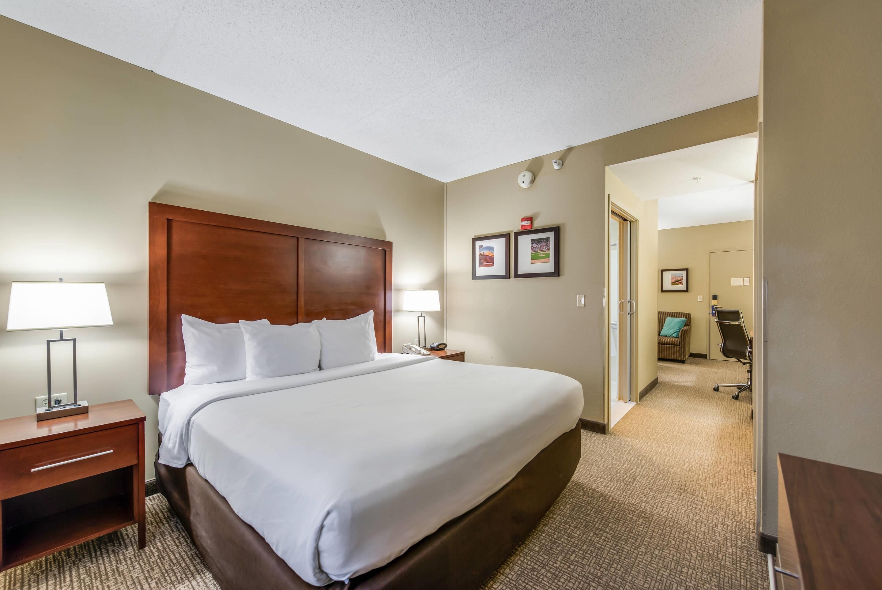 A hotel room with a large bed, wooden headboard, two bedside tables with lamps, a carpeted floor, and a hallway leading to a bathroom.