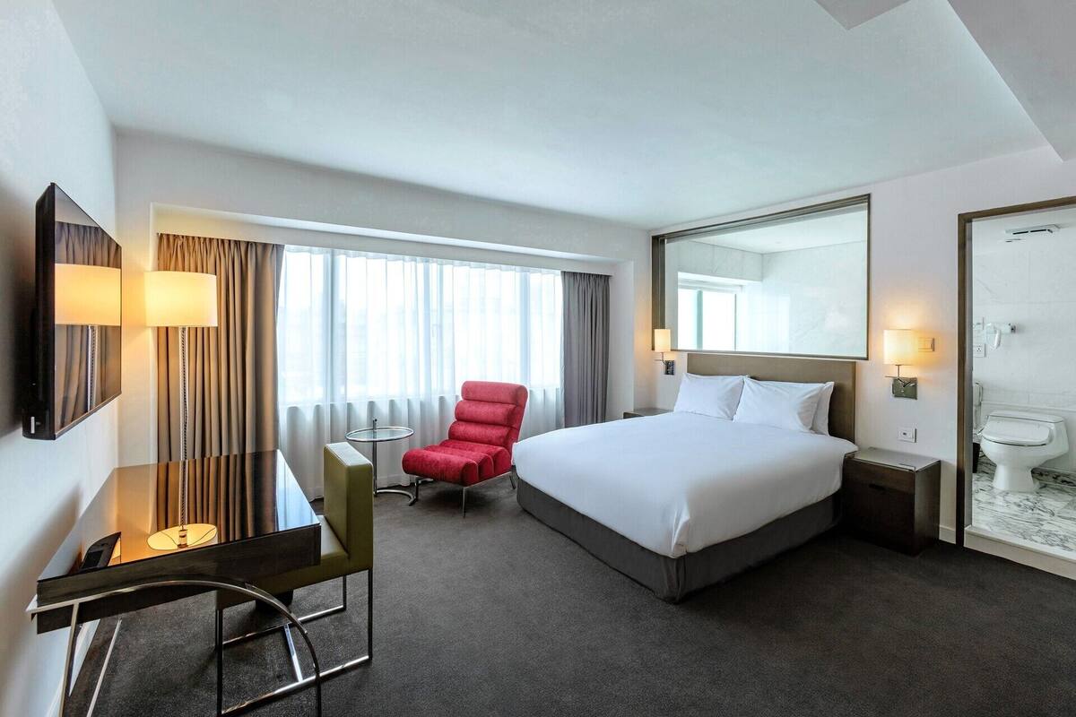 A hotel room with a bed, a desk, a chair, a round table, a mirror, and a bathroom.