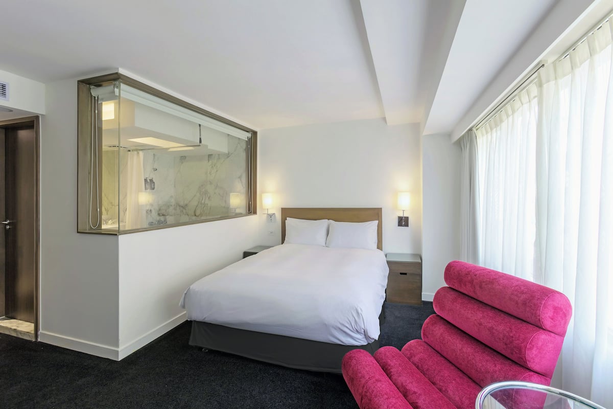 A hotel room with a large bed, a mirror, a nightstand, a chair, and a window with curtains.