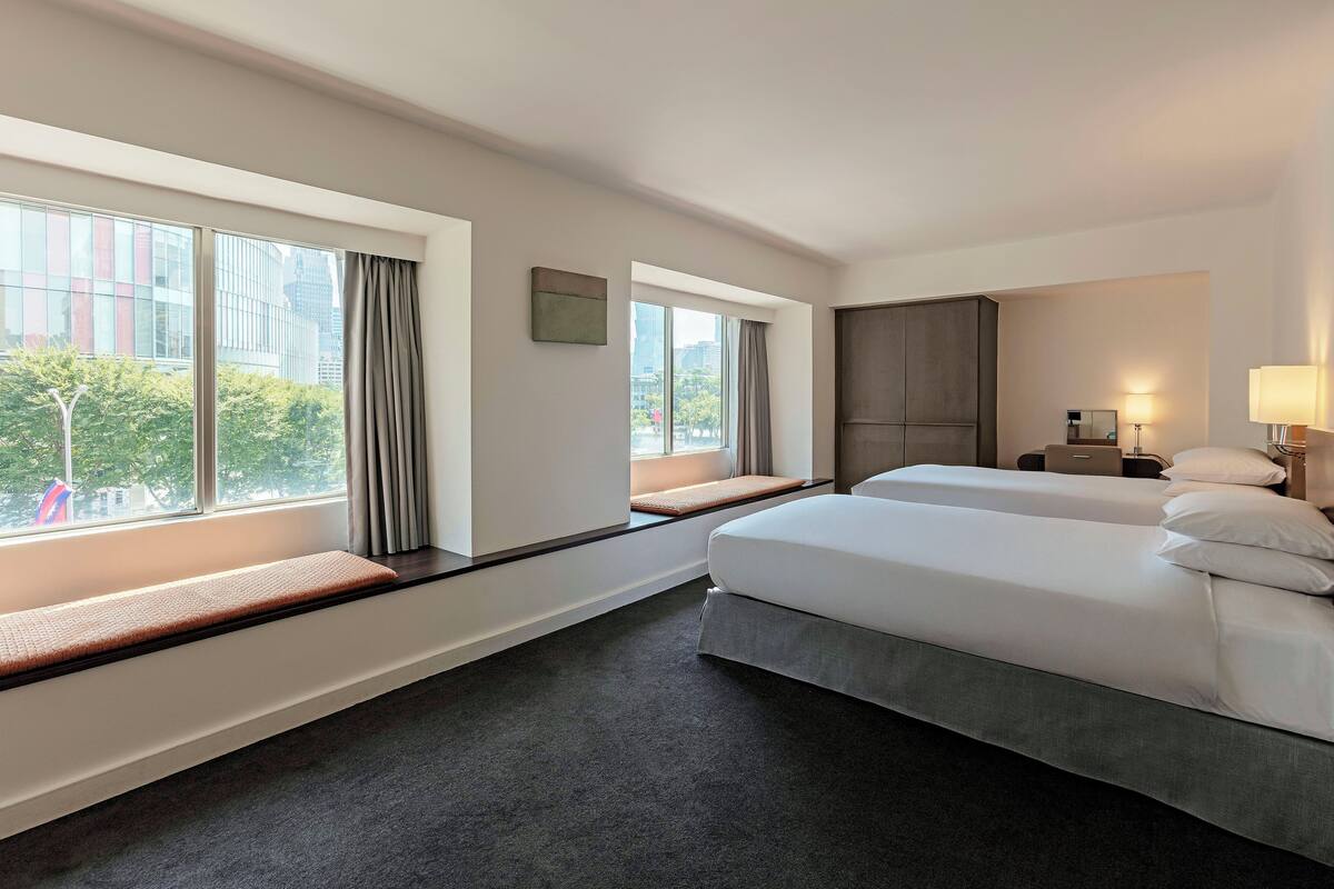 A hotel room with two beds, large windows, a bench, and a view of the city.