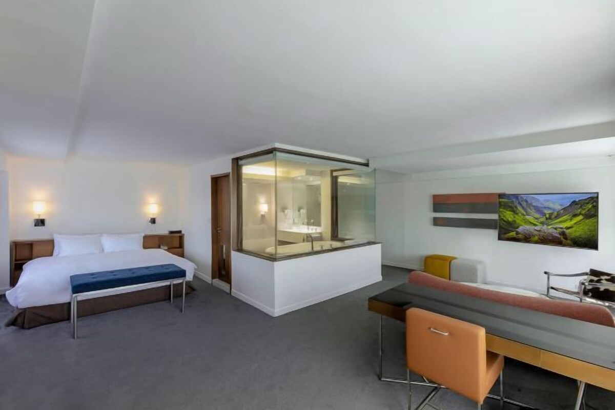 A modern hotel room with a bed, a desk, a bench, a glass-enclosed bathroom, and a painting on the wall.