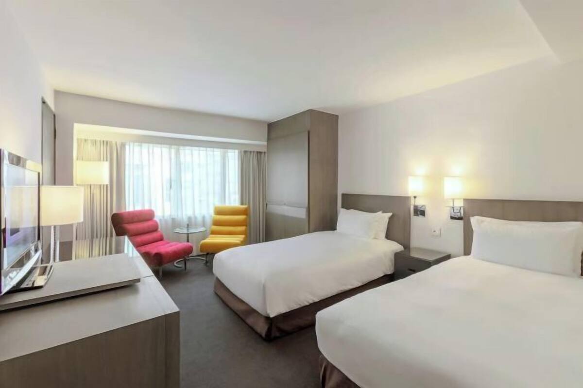 A hotel room with two beds, a desk with a laptop, a TV, a chair, a small table, and a window with curtains.