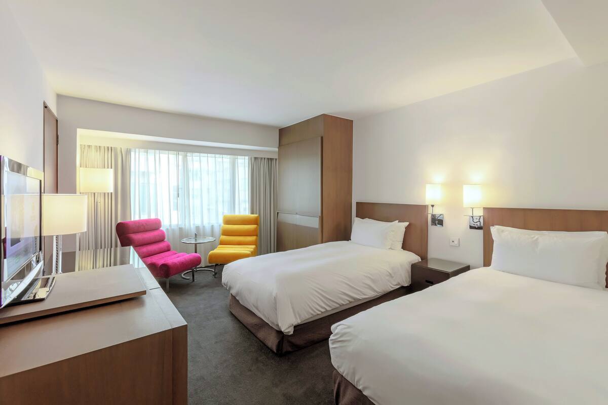 A hotel room with two beds, a desk with a laptop, a chair, a TV, and a window with curtains.