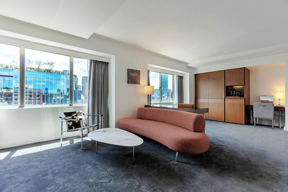 A modern hotel room with a sofa, a small table, a chair, a desk, and a kitchen area. Large windows offer a view of the city.
