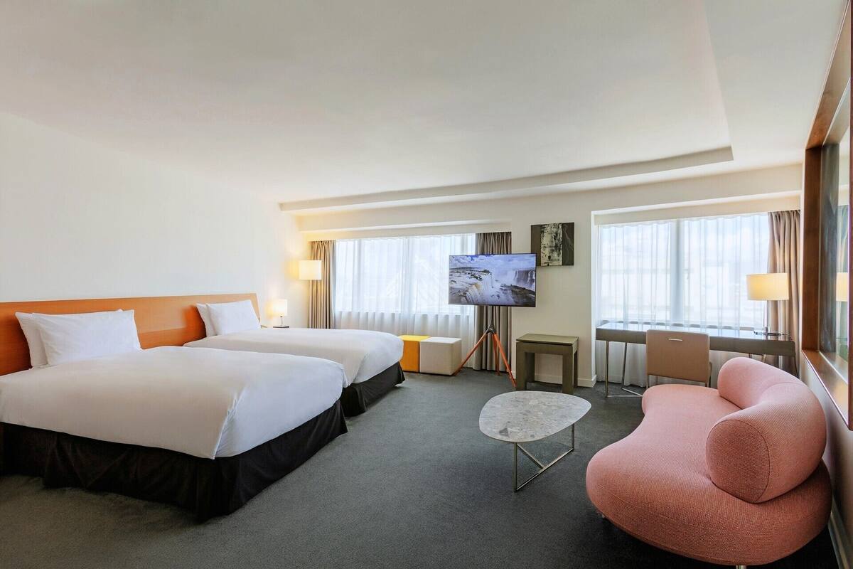 A hotel room with two beds, a television, a desk, a chair, and large windows.