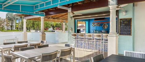 An outdoor patio area with tables and chairs, a bar with a chalkboard menu, and a view of the surrounding greenery.