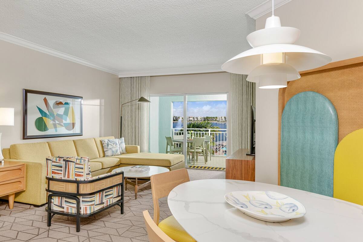 Une chambre d’hôtel moderne dotée d’un canapé, d’une table à manger, d’une table d’appoint en osier et d’un balcon avec vue.