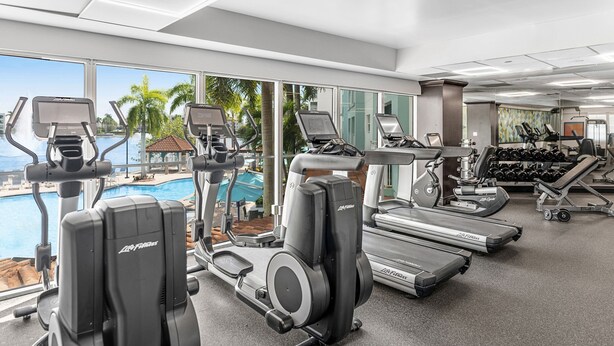 Une salle de sport équipée de tapis de course, de vélos elliptiques et d’haltères, avec vue sur une piscine bordée de palmiers.
