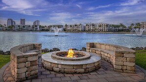 Un foyer circulaire en pierre, avec un feu au centre, entouré de bancs en pierre, et offrant une vue sur un lac avec des fontaines et des bâtiments en arrière-plan.