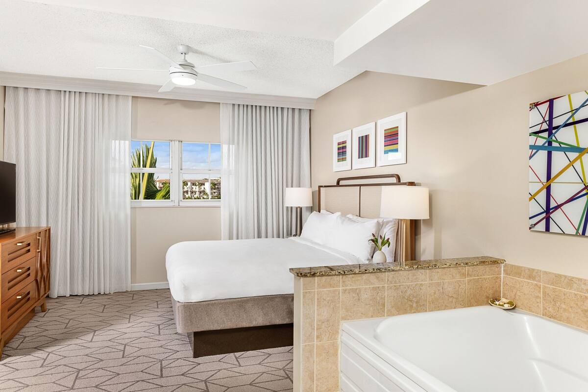 Une chambre d’hôtel comprenant un lit, un bureau, une télévision, un ventilateur de plafond, une fenêtre avec des rideaux, une salle de bain avec une baignoire et des œuvres d’art accrochées au mur.