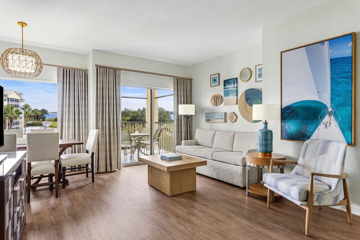 Amplia sala de estar con una mesa de centro de madera, un sofá y un sillón a rayas. Cuenta con un ventanal con cortinas, una zona de comedor con sillas y vistas a un edificio y zonas verdes.