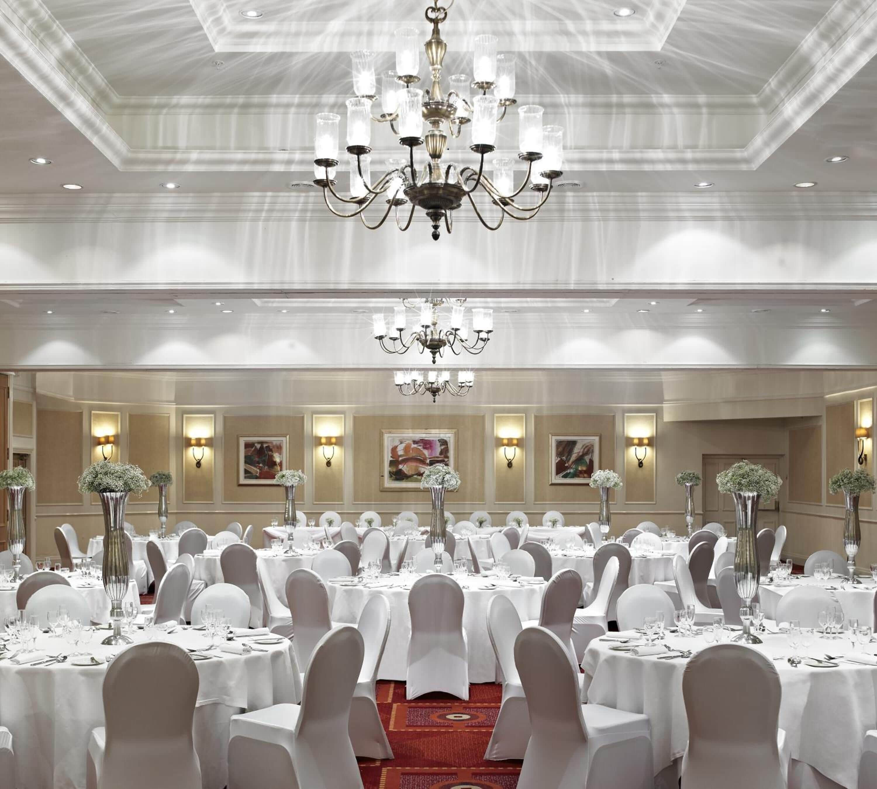 Un salón de banquetes con varias mesas redondas, manteles blancos y altos jarrones con flores.