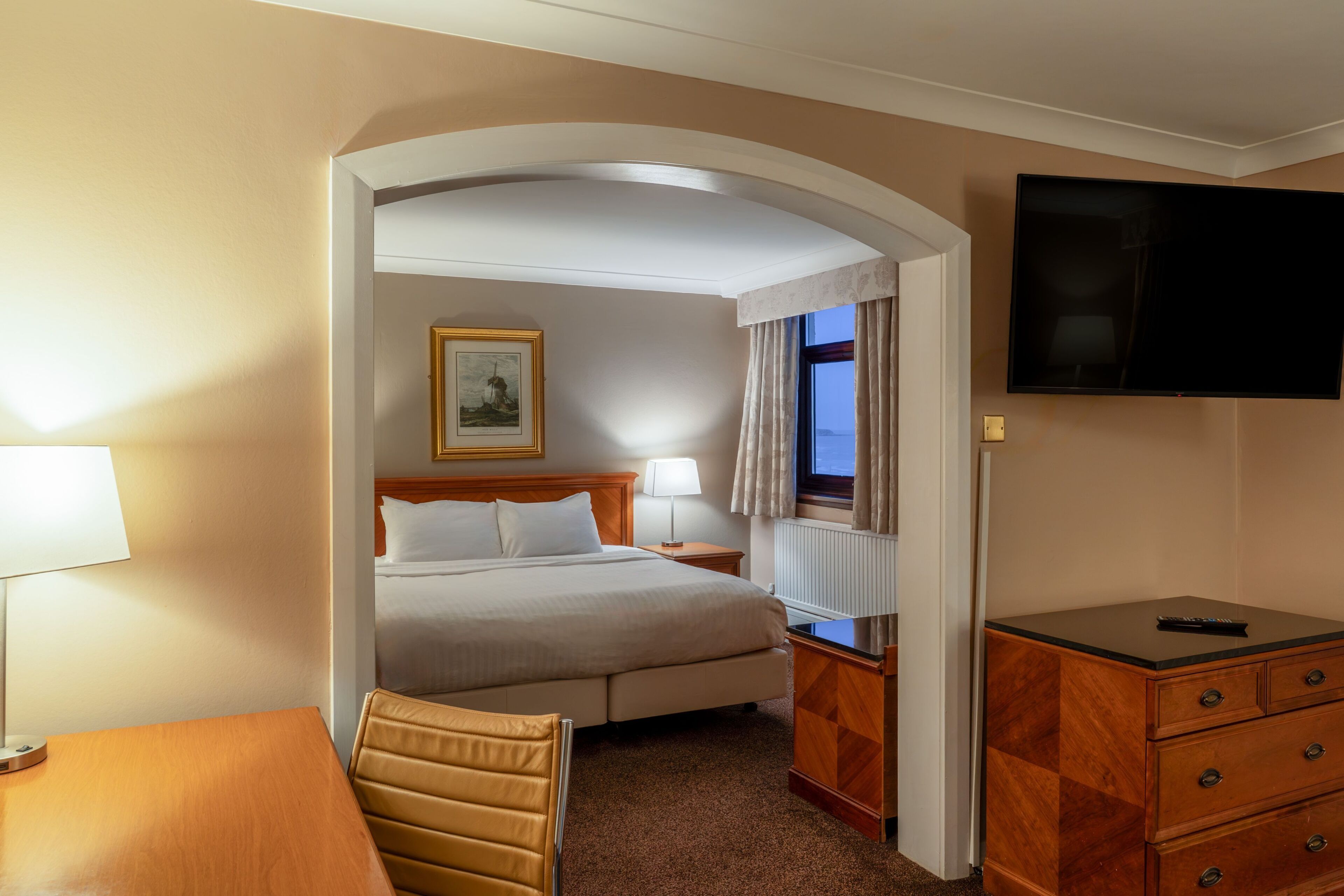 Une chambre d’hôtel comprenant un lit, un bureau, une chaise, une commode, un téléviseur et un tableau encadré au mur.