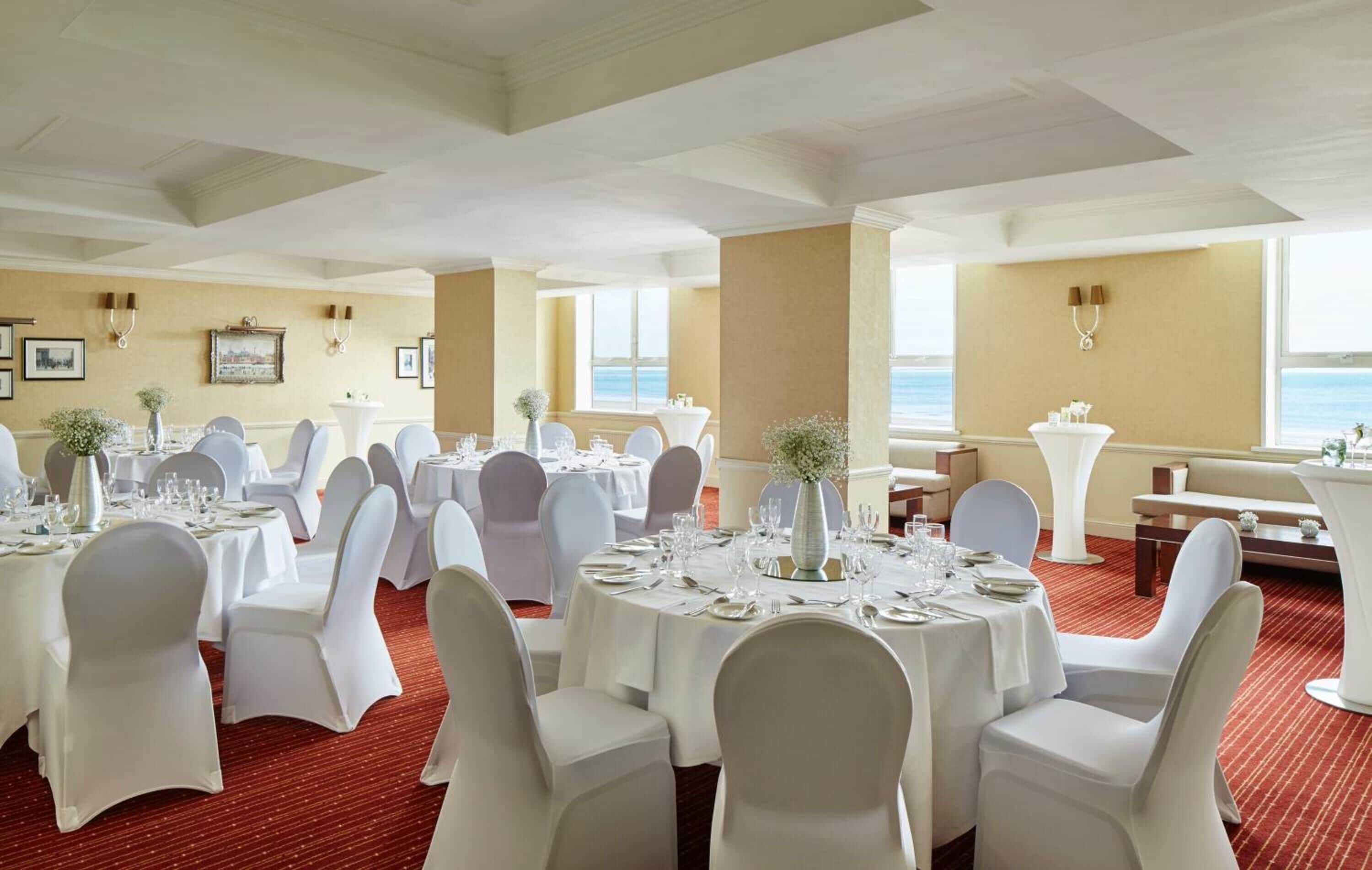 Un comedor de hotel con mesas redondas dispuestas para un evento formal, sillas blancas y vistas al mar a través de amplias ventanas.