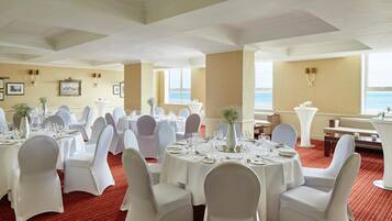 Un comedor de hotel con mesas redondas dispuestas para un evento formal, sillas blancas y vistas al mar a través de amplias ventanas.