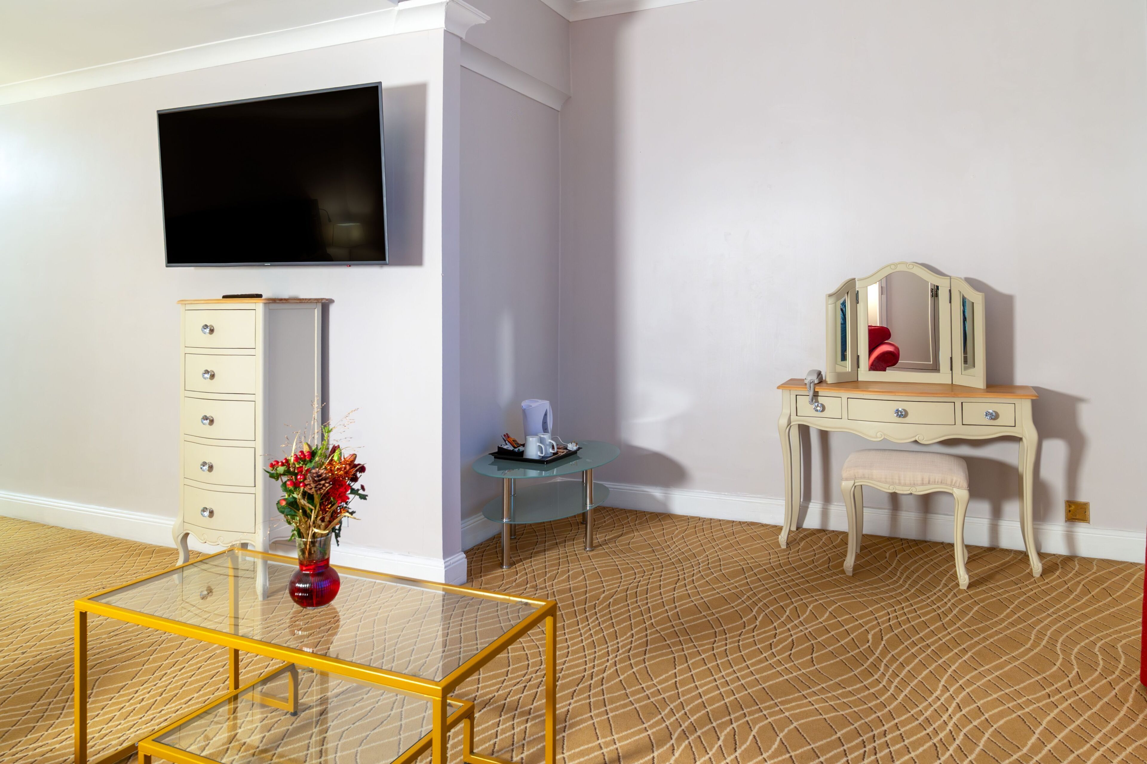 A room with a TV, a dresser, a vanity with a mirror, a small table with a vase, and a glass coffee table.