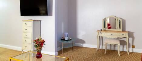 A room with a TV, a dresser, a vanity with a mirror, a small table with a vase, and a glass coffee table.