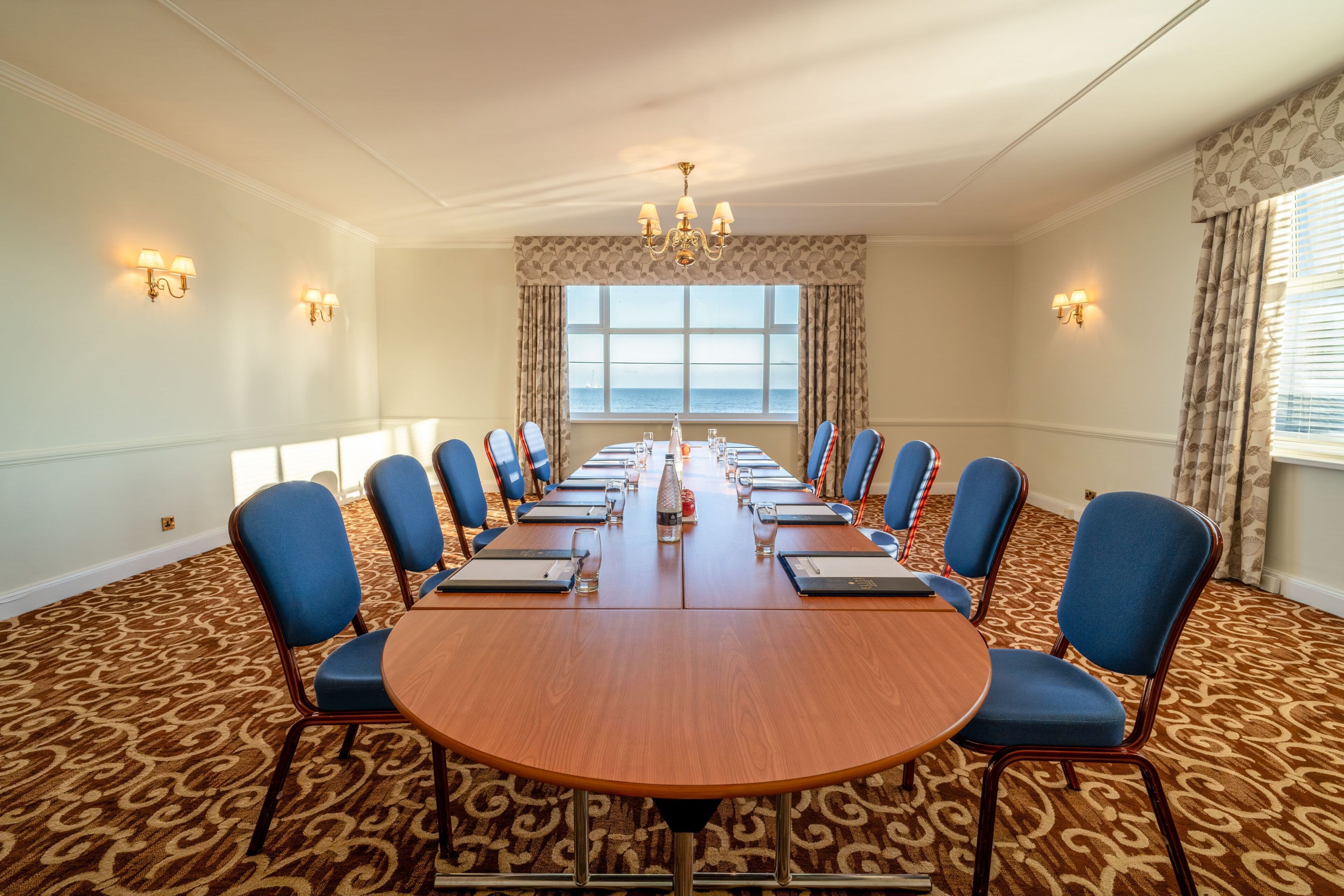 A conference room with a long table, blue chairs, and a view of the sea through large windows.