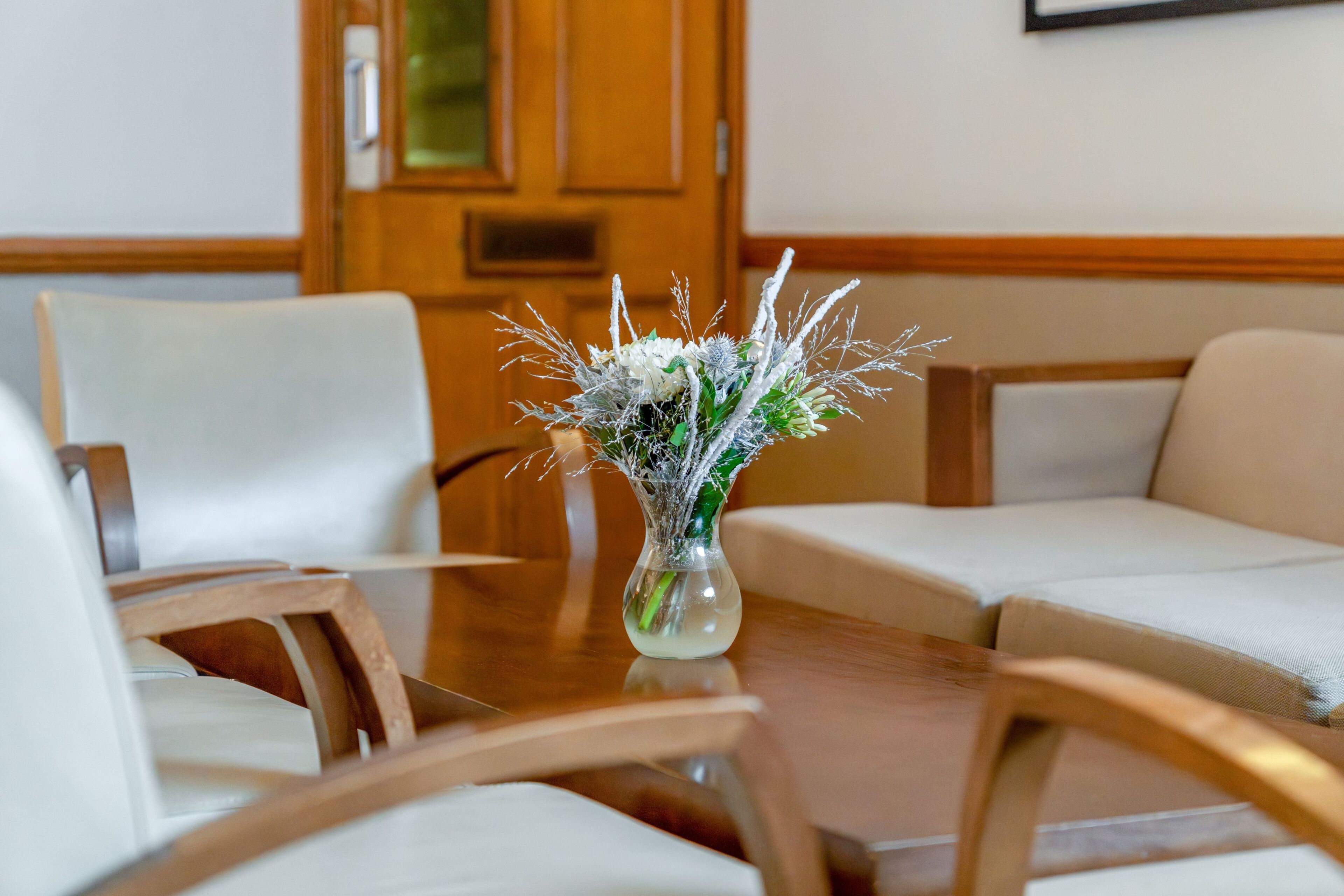 A wooden table with a clear glass vase containing white flowers and greenery.