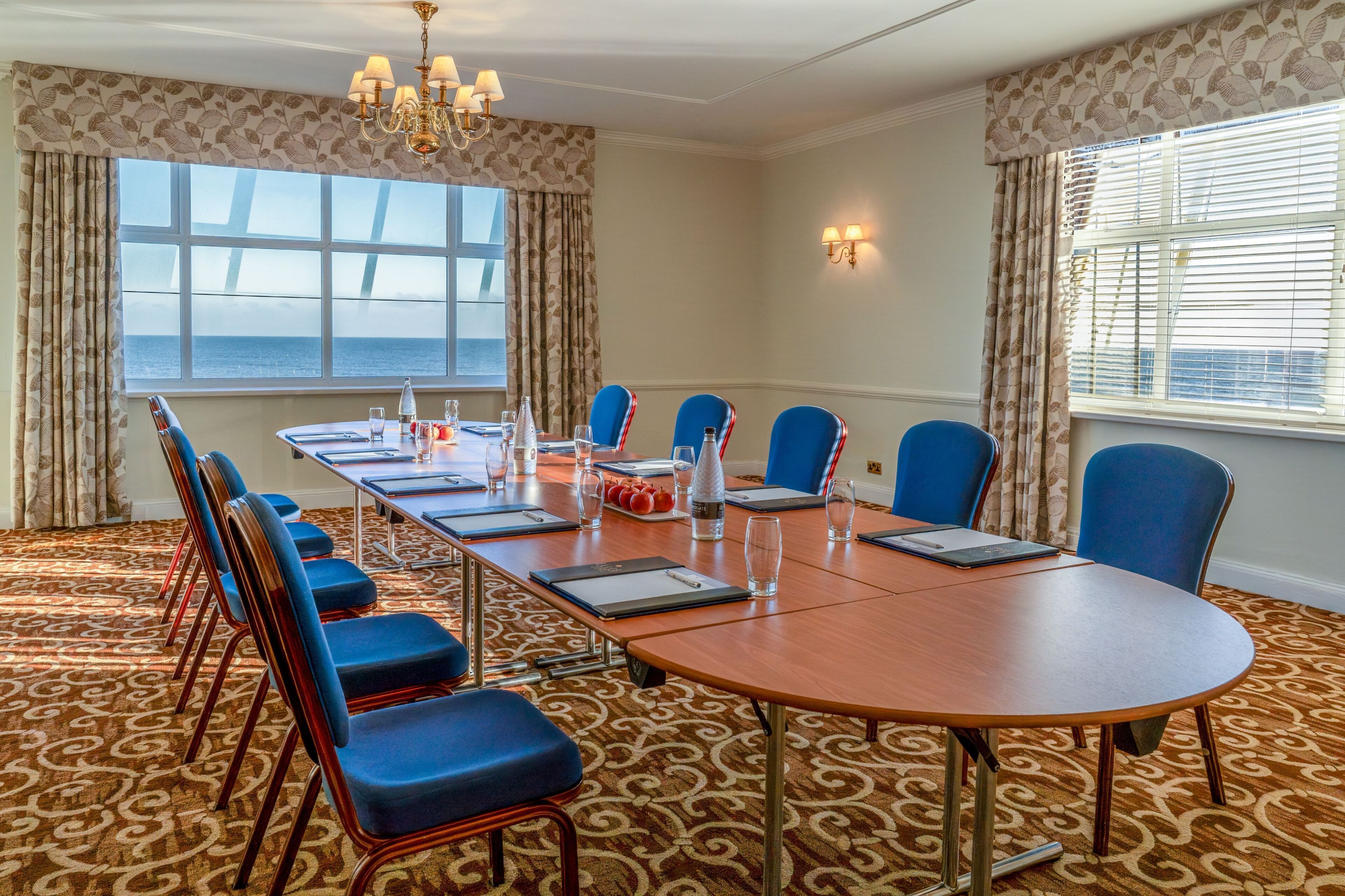 Un salón de conferencias con una larga mesa, sillas azules, una araña y vistas al mar a través de amplias ventanas.