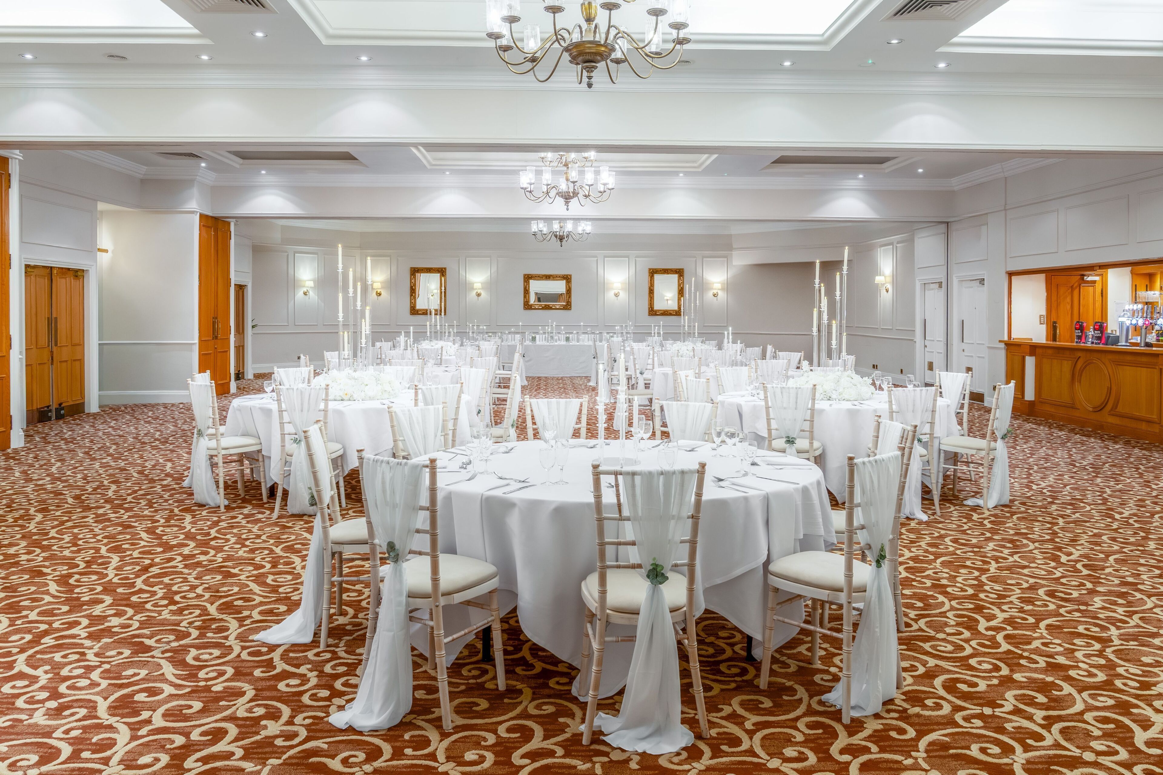 Une grande salle de banquet avec des tables rondes, des nappes blanches et des chaises ornées de rubans blancs.