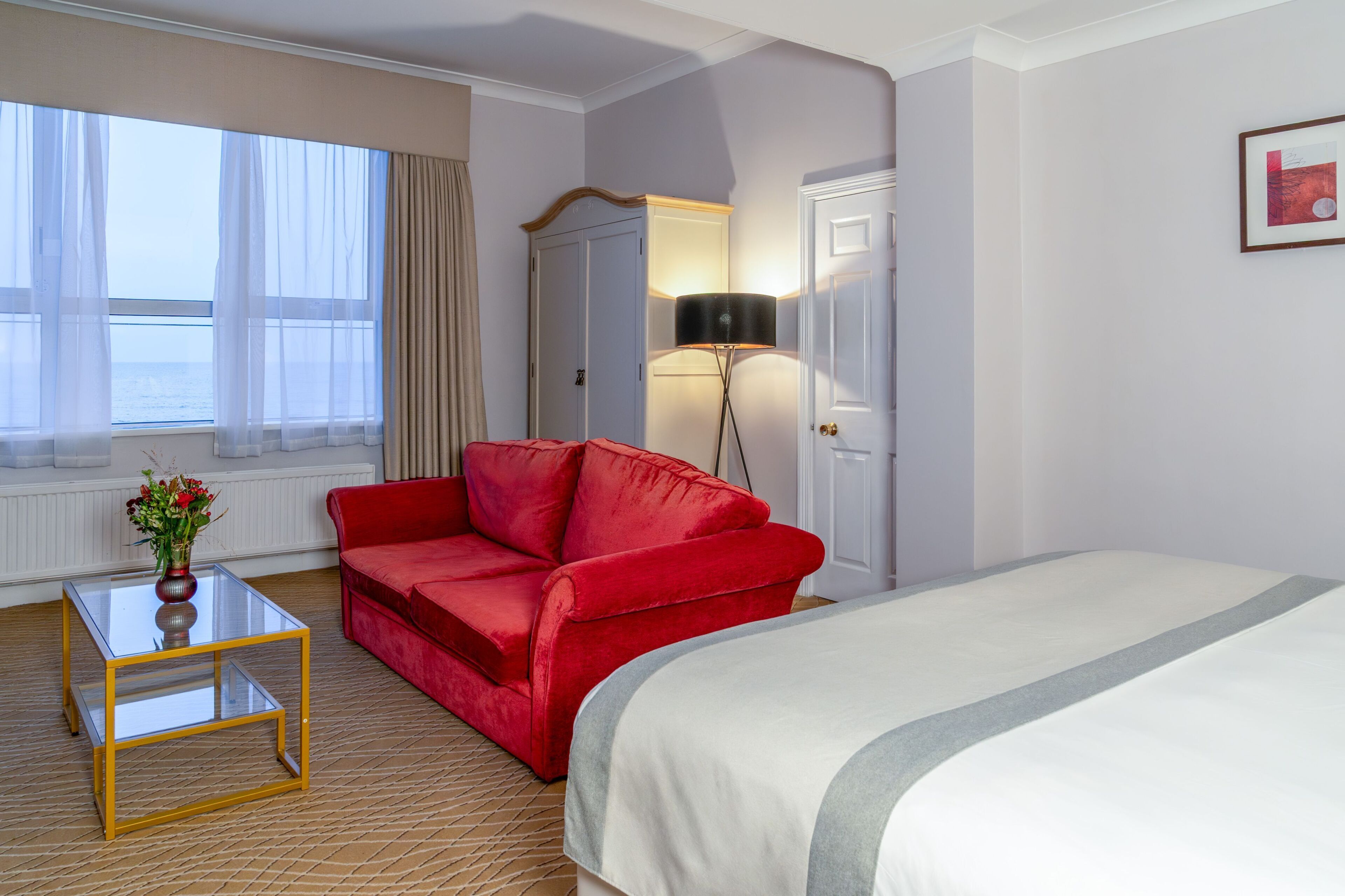 Hotellihuone, jossa on sänky, punainen sohva, lasitasoinen pöytä, jossa on maljakko, lamppu ja näkymä merelle läpikuultavien verhojen läpi.