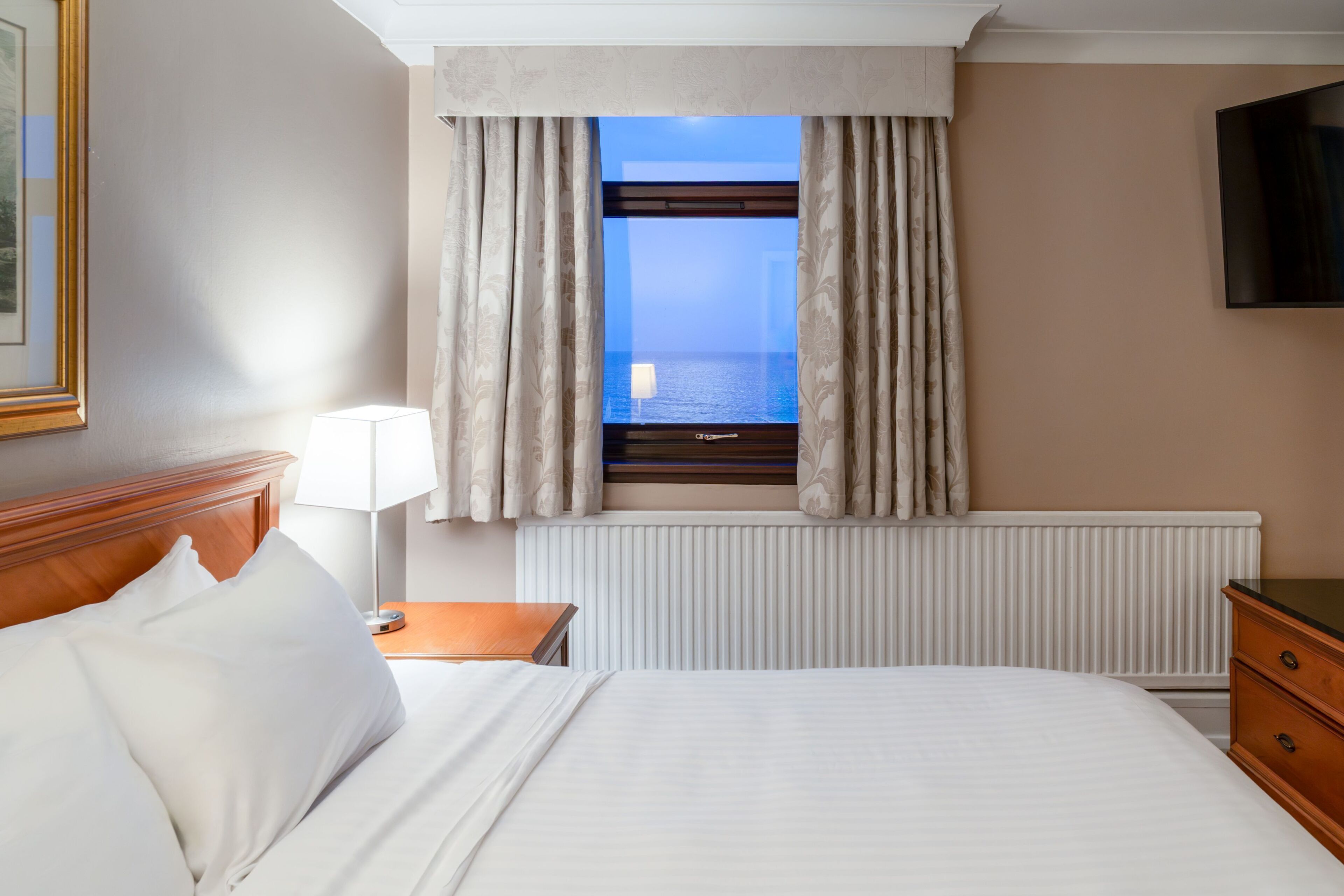 Habitación de hotel con cama, mesita de noche, lámpara, cómoda, ventana con cortinas y televisor en la pared.